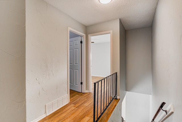 corridor with light wood-style floors, a textured ceiling, baseboards, an upstairs landing, and visible vents