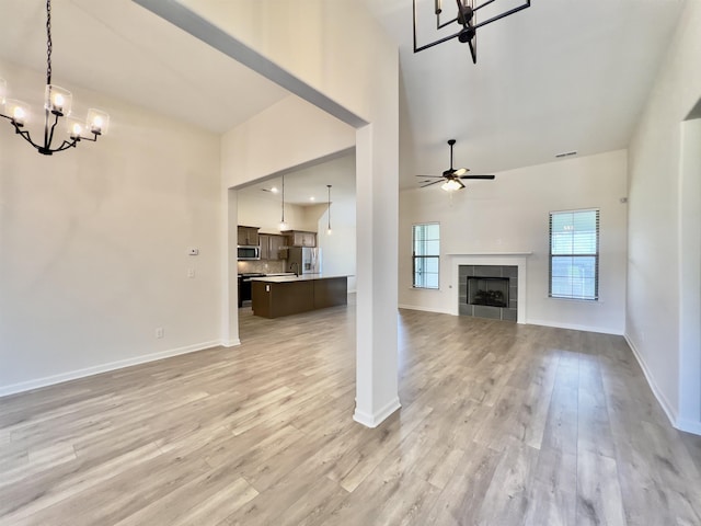 unfurnished living room with a towering ceiling, baseboards, a tiled fireplace, light wood-style flooring, and ceiling fan with notable chandelier
