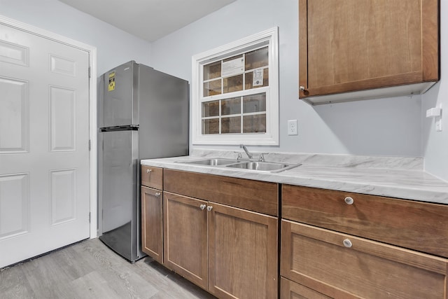 kitchen featuring freestanding refrigerator, brown cabinets, light stone counters, a sink, and light wood-style flooring