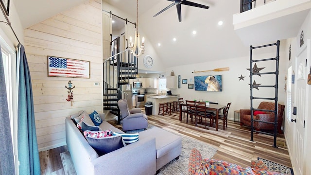 living room featuring stairs, wood finished floors, wooden walls, high vaulted ceiling, and ceiling fan with notable chandelier