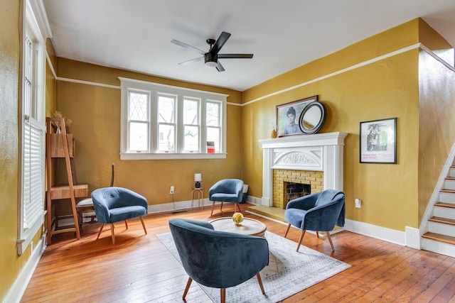 living area featuring wood-type flooring, stairs, a brick fireplace, baseboards, and a ceiling fan