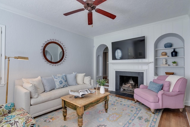 living area with ornamental molding, a fireplace with flush hearth, wood finished floors, a textured ceiling, and a ceiling fan