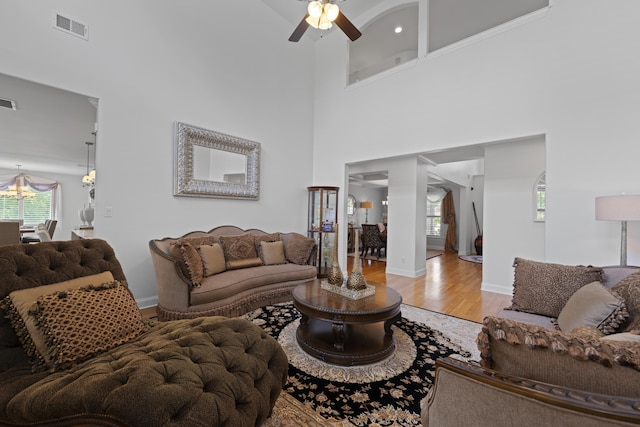 living room featuring visible vents, baseboards, a high ceiling, light wood-style flooring, and ceiling fan with notable chandelier