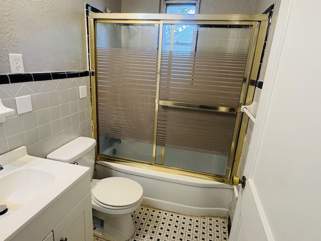 bathroom with tile walls, bath / shower combo with glass door, vanity, toilet, and a textured wall
