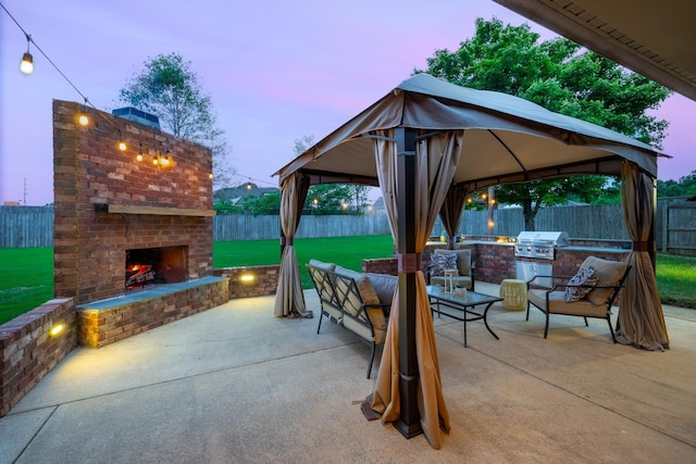 patio terrace at dusk featuring exterior kitchen, a gazebo, a fenced backyard, an outdoor brick fireplace, and a grill
