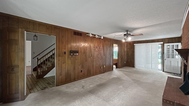 unfurnished living room featuring a textured ceiling, wood walls, stairway, ceiling fan, and light colored carpet