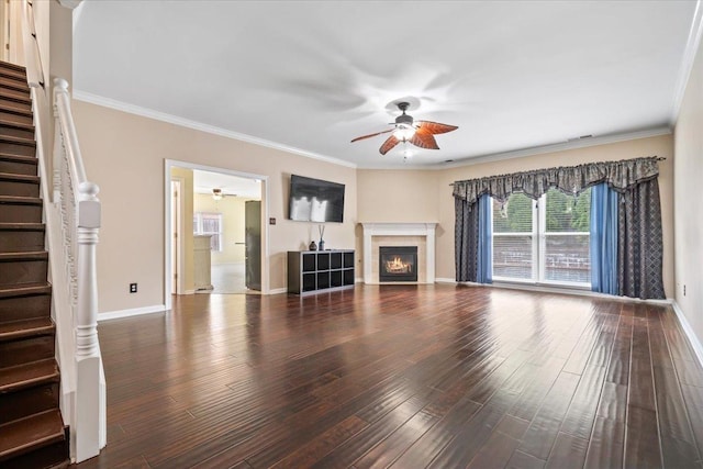 unfurnished living room with crown molding, stairs, a ceiling fan, baseboards, and a lit fireplace
