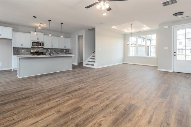 unfurnished living room with a tray ceiling, dark wood-style flooring, visible vents, a sink, and ceiling fan with notable chandelier