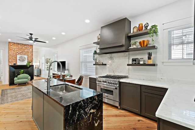 kitchen with light stone countertops, high end stove, a sink, open shelves, and wall chimney exhaust hood