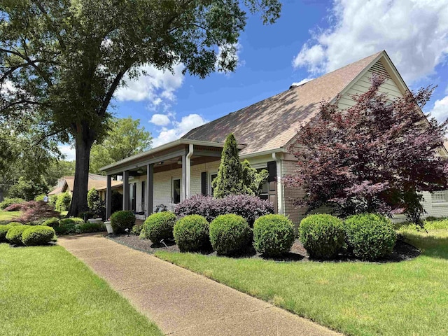 view of side of property with a lawn, roof with shingles, brick siding, and covered porch