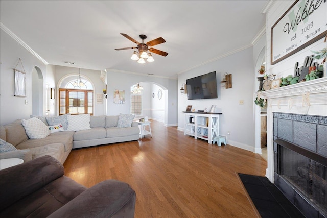 living room with arched walkways, a tiled fireplace, a ceiling fan, wood finished floors, and crown molding
