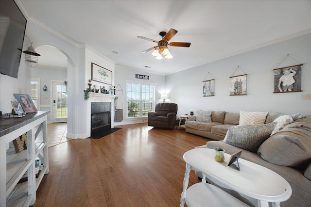 living area featuring ceiling fan, light wood-style flooring, arched walkways, crown molding, and a fireplace with flush hearth