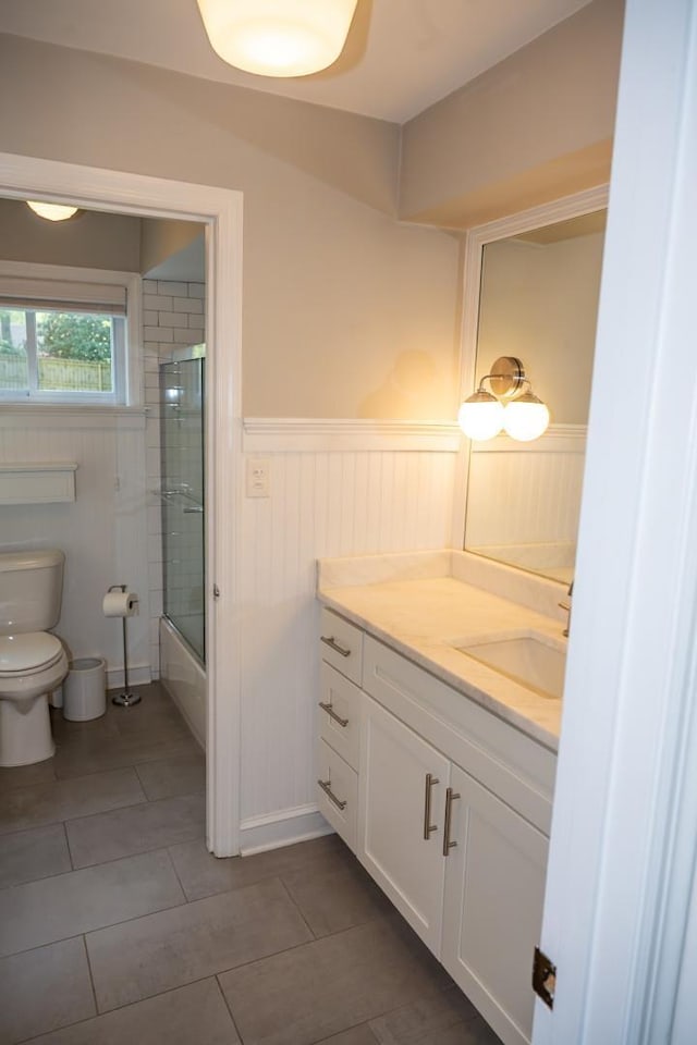 bathroom with toilet, a wainscoted wall, vanity, tile patterned flooring, and shower / bath combination with glass door