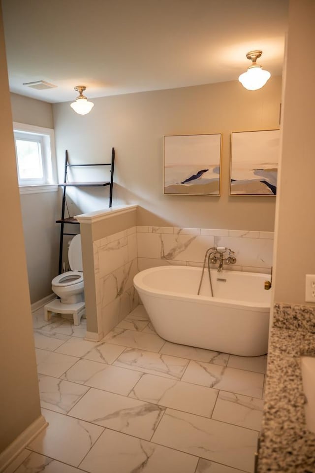 full bath featuring marble look tiles, a freestanding bath, wainscoting, toilet, and tile walls