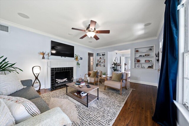 living area with built in shelves, ceiling fan, wood finished floors, crown molding, and a tiled fireplace