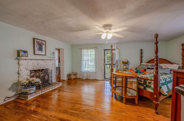 bedroom with a textured ceiling, wood finished floors, access to exterior, a stone fireplace, and ceiling fan