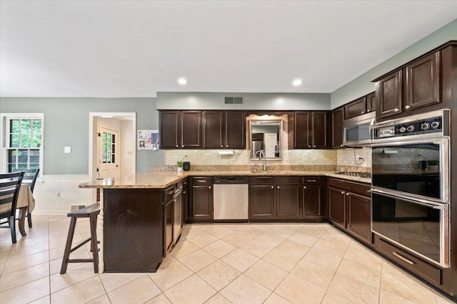 kitchen featuring appliances with stainless steel finishes, dark brown cabinetry, a peninsula, a wainscoted wall, and a sink