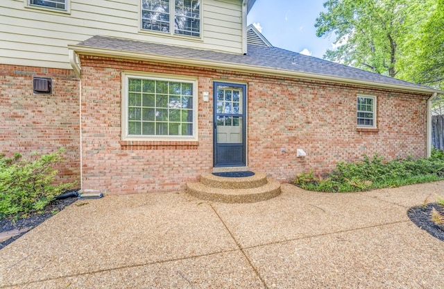 view of exterior entry featuring roof with shingles, brick siding, and a patio area