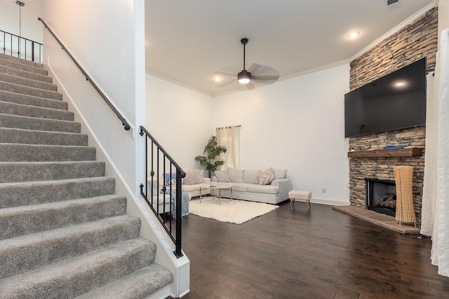 living area featuring a ceiling fan, stairs, wood finished floors, crown molding, and a stone fireplace