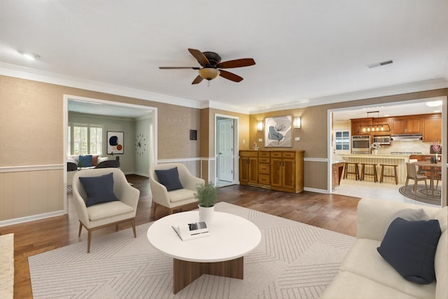 living area featuring dark wood-style flooring, crown molding, a ceiling fan, and wainscoting