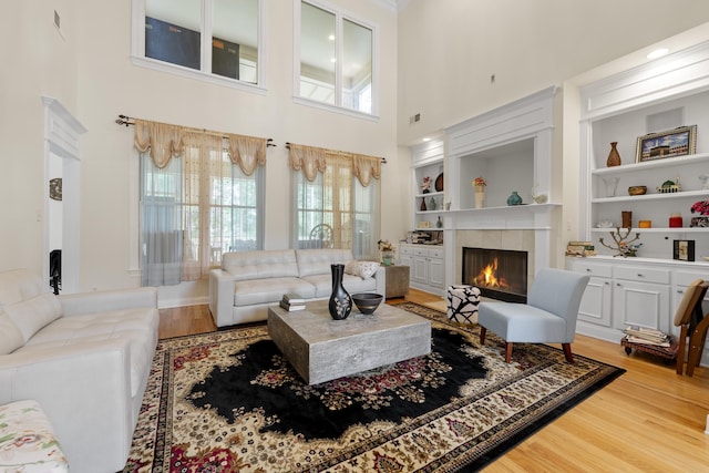 living room featuring light wood-style floors, a tile fireplace, plenty of natural light, a towering ceiling, and built in shelves