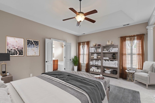 bedroom featuring light carpet, a tray ceiling, ensuite bathroom, a ceiling fan, and ornate columns
