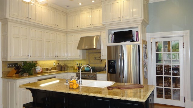 kitchen featuring backsplash, stainless steel fridge with ice dispenser, a center island with sink, light stone counters, and wall chimney exhaust hood