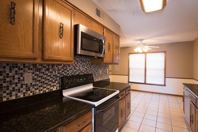 kitchen featuring light tile flooring, ceiling fan, appliances with stainless steel finishes, backsplash, and dark stone countertops