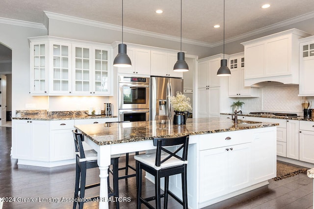 kitchen with dark hardwood / wood-style floors, dark stone counters, decorative light fixtures, a center island with sink, and appliances with stainless steel finishes