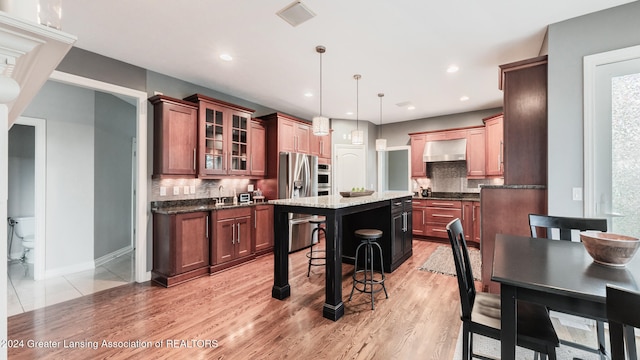 kitchen featuring a breakfast bar area, a kitchen island with sink, wall chimney range hood, dark stone countertops, and glass insert cabinets