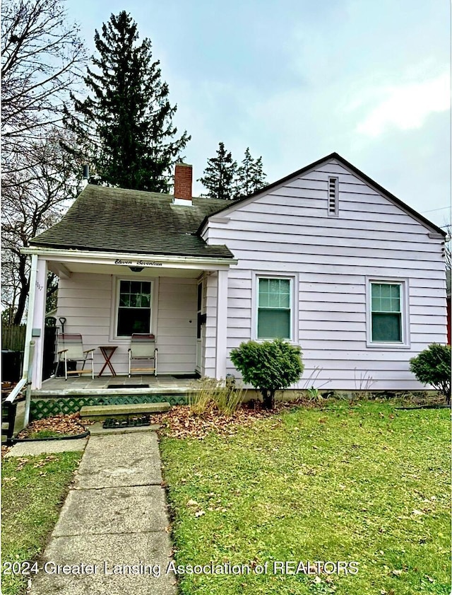 view of front of house featuring a chimney, covered porch, a front lawn, and a shingled roof