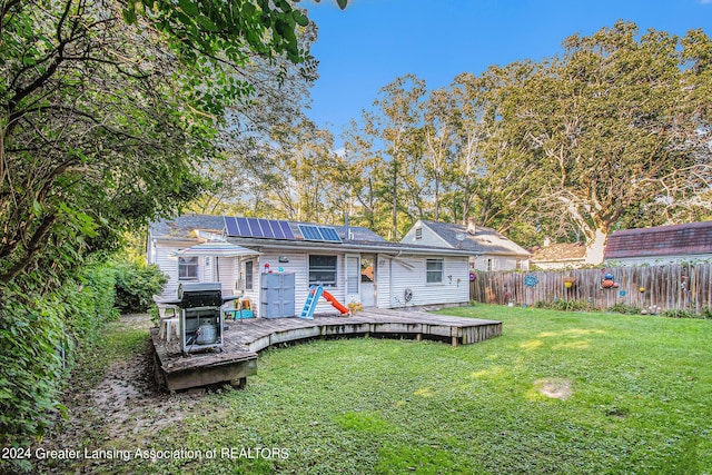 back of property featuring a wooden deck, roof mounted solar panels, a lawn, and fence