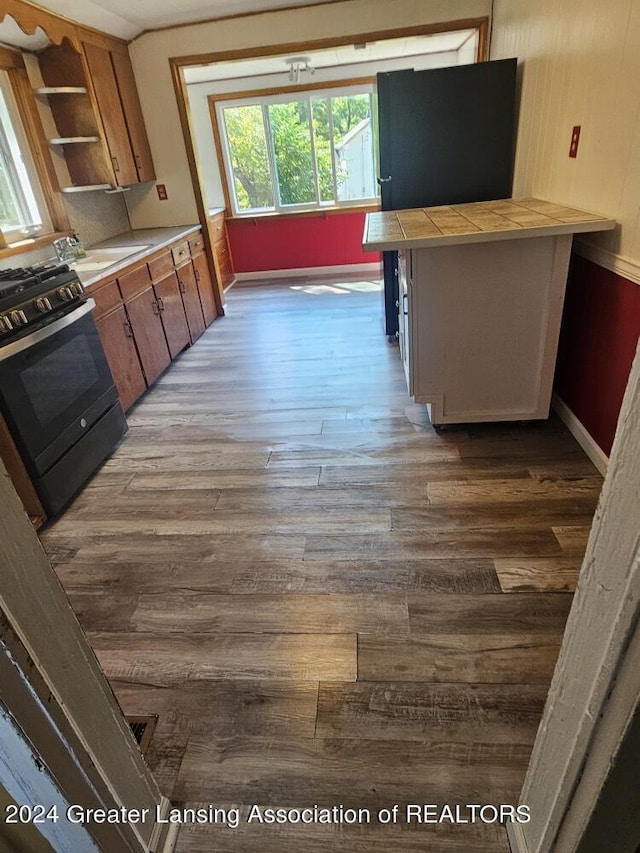 kitchen with wood finished floors, tile counters, open shelves, black gas range, and brown cabinets