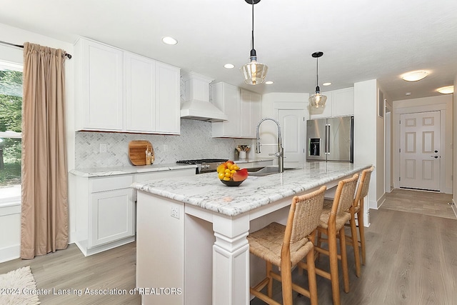 kitchen featuring premium range hood, light hardwood / wood-style floors, a center island with sink, high end refrigerator, and white cabinets