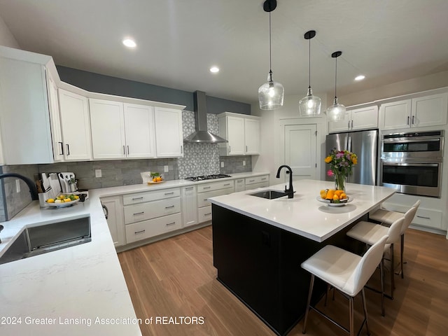 kitchen featuring sink, white cabinets, an island with sink, wall chimney range hood, and appliances with stainless steel finishes