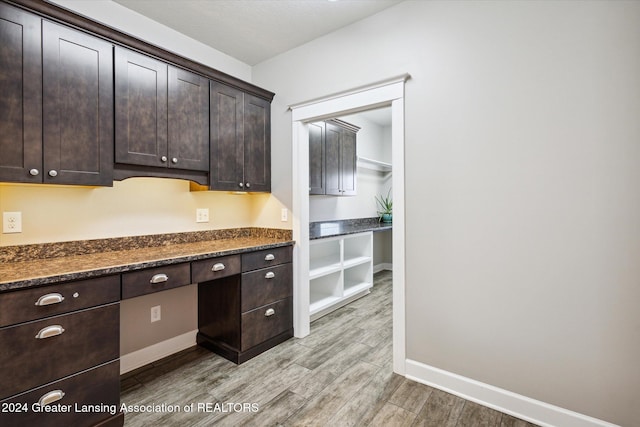 kitchen with dark brown cabinets, dark stone countertops, built in desk, and light hardwood / wood-style flooring