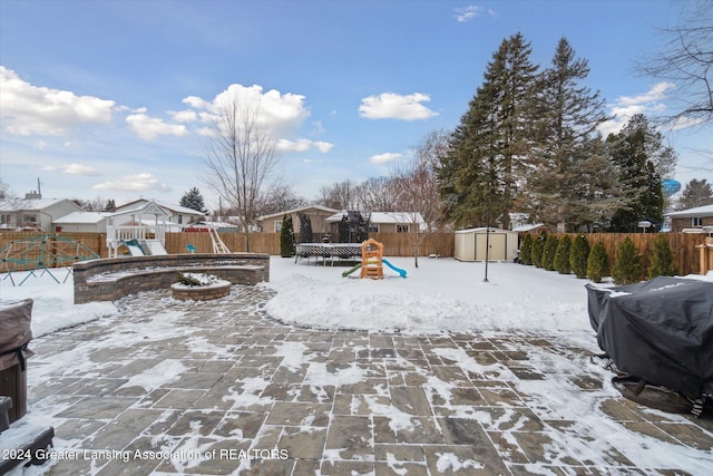 snow covered patio with a fire pit, a trampoline, a storage shed, a playground, and a grill