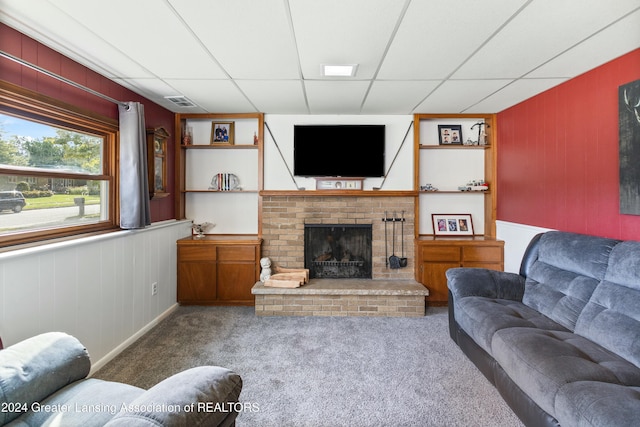 carpeted living room featuring a brick fireplace, a drop ceiling, and built in shelves