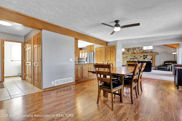 dining area featuring ceiling fan, a stone fireplace, light hardwood / wood-style flooring, and a textured ceiling