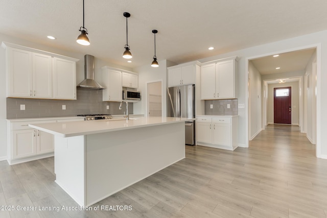 kitchen with an island with sink, white cabinets, hanging light fixtures, stainless steel appliances, and wall chimney range hood