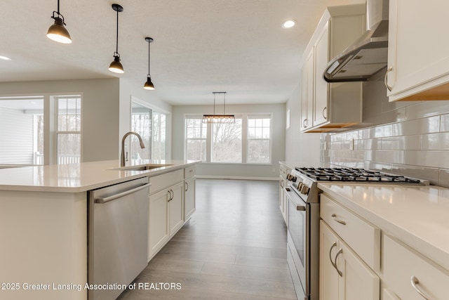 kitchen with sink, hanging light fixtures, appliances with stainless steel finishes, an island with sink, and wall chimney range hood