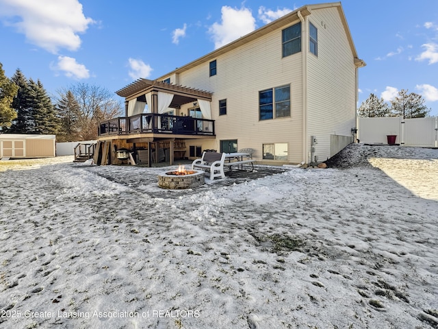 snow covered back of property with a wooden deck, a pergola, a shed, and a fire pit