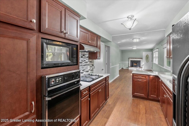 kitchen featuring black appliances, under cabinet range hood, open floor plan, light wood-style floors, and a stone fireplace