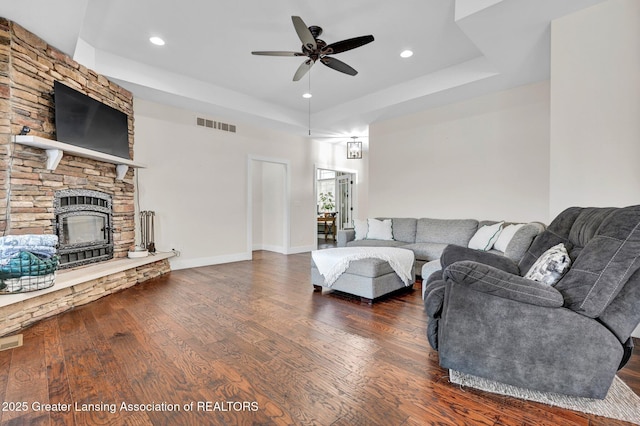 living room featuring a stone fireplace, dark hardwood / wood-style floors, ceiling fan, and a tray ceiling