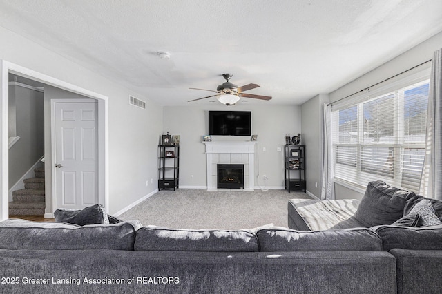 carpeted living area featuring baseboards, visible vents, a ceiling fan, a tiled fireplace, and stairs