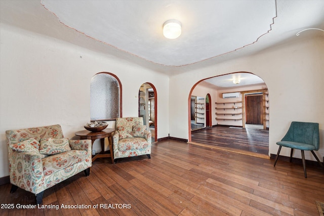 sitting room with arched walkways, an AC wall unit, baseboards, and wood finished floors