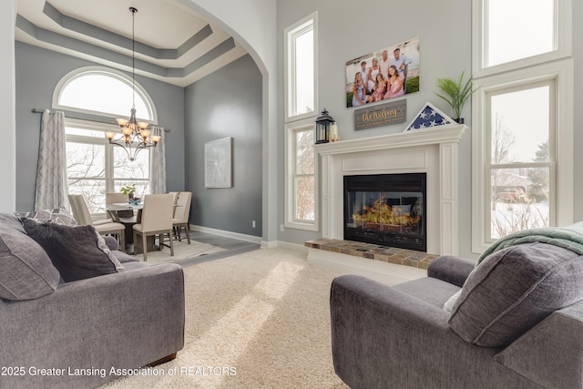 carpeted living room featuring a notable chandelier, a high ceiling, a glass covered fireplace, baseboards, and a raised ceiling