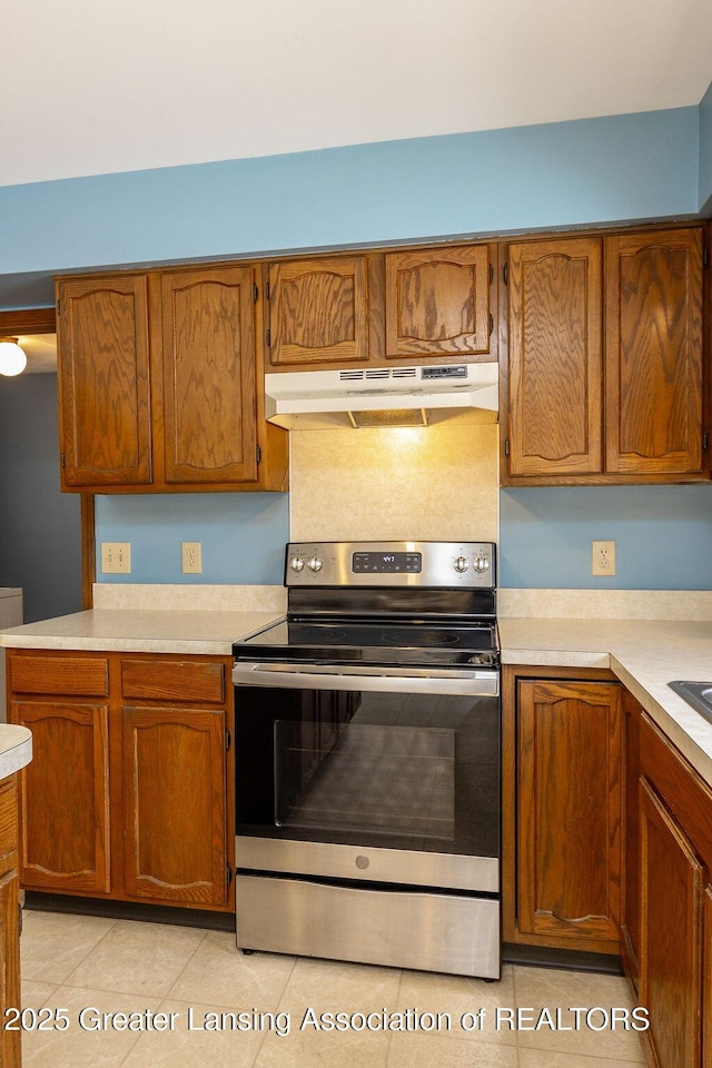 kitchen with light tile patterned floors, under cabinet range hood, electric range, light countertops, and brown cabinets