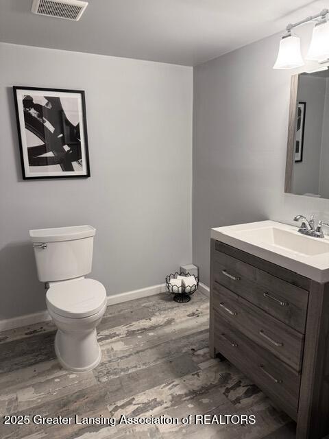 half bath with visible vents, toilet, vanity, wood finished floors, and baseboards