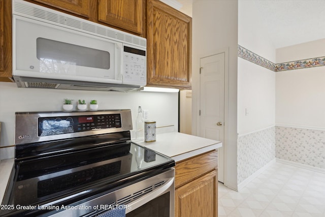 kitchen with white microwave, a wainscoted wall, brown cabinets, light countertops, and stainless steel range with electric cooktop